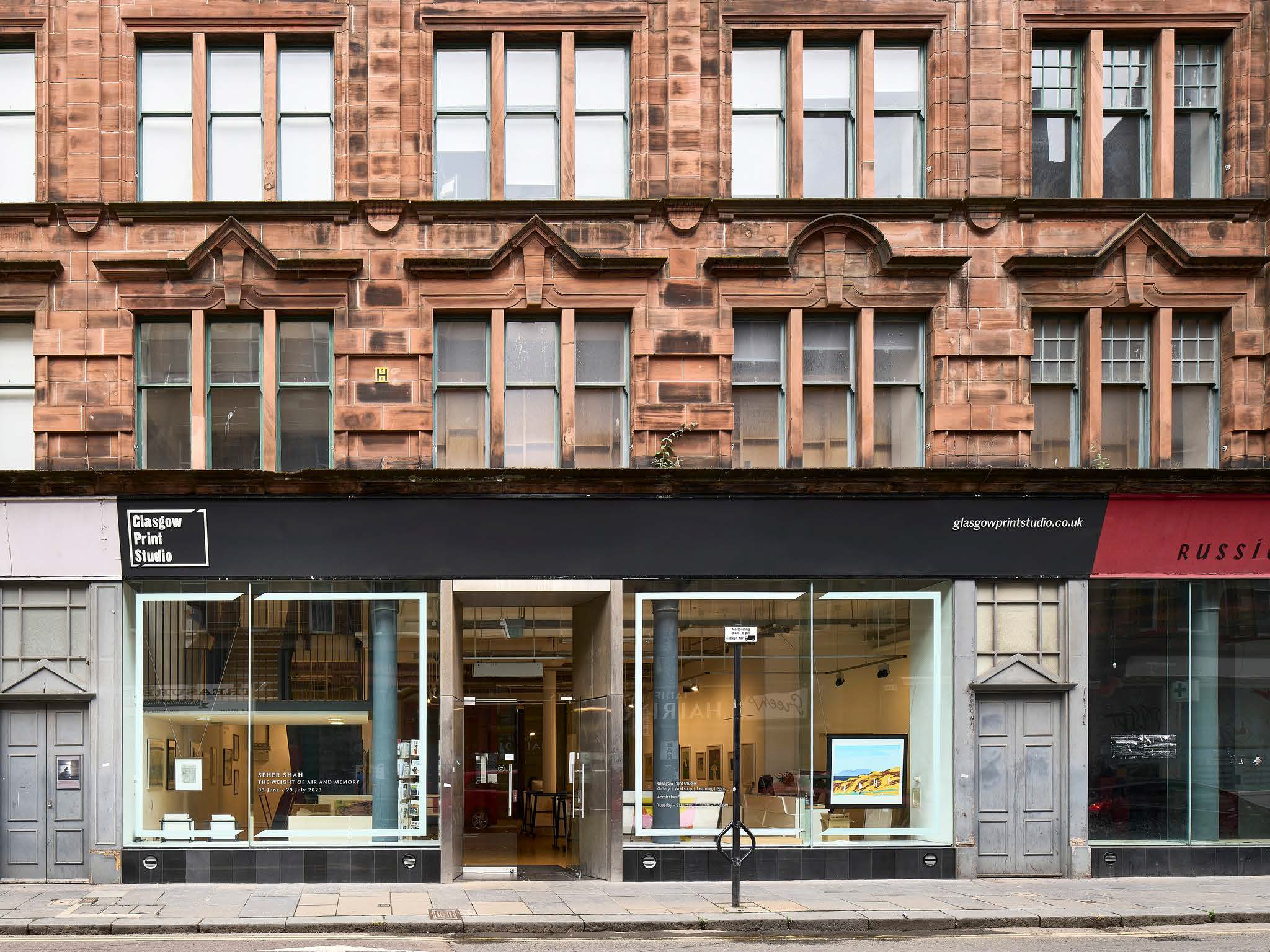 GPS Shop Exterior The image shows the exterior of the Glasgow Print Studio, housed in a red sandstone building with large windows and modern black signage. The studio's display windows reveal framed artworks and a glimpse into the bright gallery space inside.
